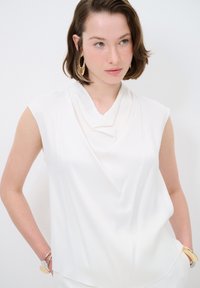 Top bianco senza maniche con scollatura drappeggiata, texture liscia e vestibilità morbida. Il modello indossa orecchini a cerchio dorati e un bracciale.