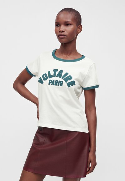 Γυναίκα με κοντά μαλλιά που φοράει λευκό t-shirt με το κείμενο "Voltaire Paris" και μπορντό δερμάτινη φούστα, στέκεται με το χέρι στη μέση.