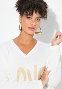Maglione bianco a V con lettering beige testurizzato e polsini a costine, abbinato a grandi orecchini geometrici dorati. Tessuto leggero e liscio.
