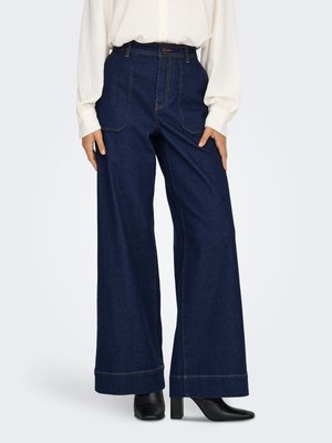 Vrouw draagt donkerblauwe high-waist wijde jeans, witte blouse met lange mouwen en zwarte enkellaarsjes met hak.