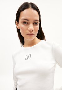 Weißes, geripptes Langarmshirt mit kleinem schwarzen, gestickten Logo auf der Brust. Die glatte Textur und das figurbetonte Design werten das Erscheinungsbild auf.