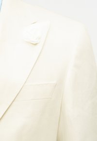 Κοντινό πλάνο για το πέτο ενός μπεζ σακακιού με μια καρφίτσα λευκού υφασμάτινου λουλουδιού και μια τσέπη στο στήθος σε ύφασμα με λεία υφή.