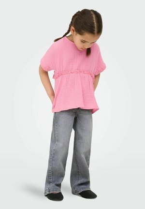 Nuori tyttö, jolla on letitetyt hiukset ja päällään vaaleanpunainen lyhythihainen paita ja löysät harmaat farkut, katsoo alas kädet takataskuissa.