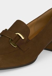 Mocassin femme en daim marron avec un petit talon carré et un détail de boucle métallique dorée sur le dessus.