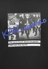 Camiseta de algodón negra con una impresión de foto vintage en gris y texto azul que dice "KARL LAGERFELD" con la cita debajo en blanco.