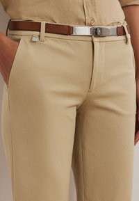 Beiga byxor av mjukt tyg med fickor i sidan, kombinerade med ett brunt läderbälte med ett silverfärgat spänne.