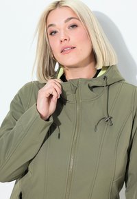 Olivově zelená softshellová bunda s kapucí, se zapínáním na přední straně na zip, akcenty na šňůrkách a hladkou texturou.