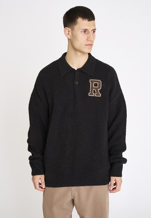 Ung mand iført en sort langærmet sweater med krave og et stort "R" på brystet, kombineret med beige bukser, ser mod højre.