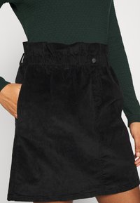 Svart manchesterkjol med hög, rynkad midjeband, sidofickor och en mjuk textur, som går till mitten av låret med rak skärning.