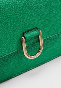 Grön texturerad läderhandväska med detaljer i guldfärgad D-ring och svart sömnad längs kanterna.