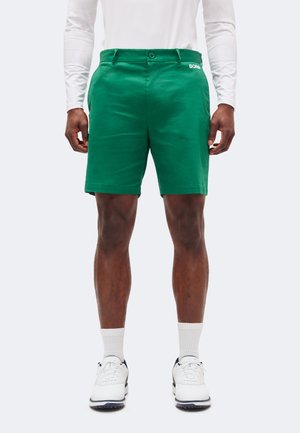 Άντρας που φοράει πράσινα σορτς, λευκό πουκάμισο με μακριά μανίκια, λευκές κάλτσες και λευκά αθλητικά παπούτσια, στέκεται με τα χέρια χαλαρά δίπλα στο σώμα.