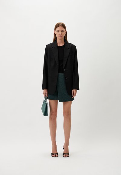 Black tailored blazer over a black top, paired with a textured green skirt and holding a small teal bag; black open-toe heels.