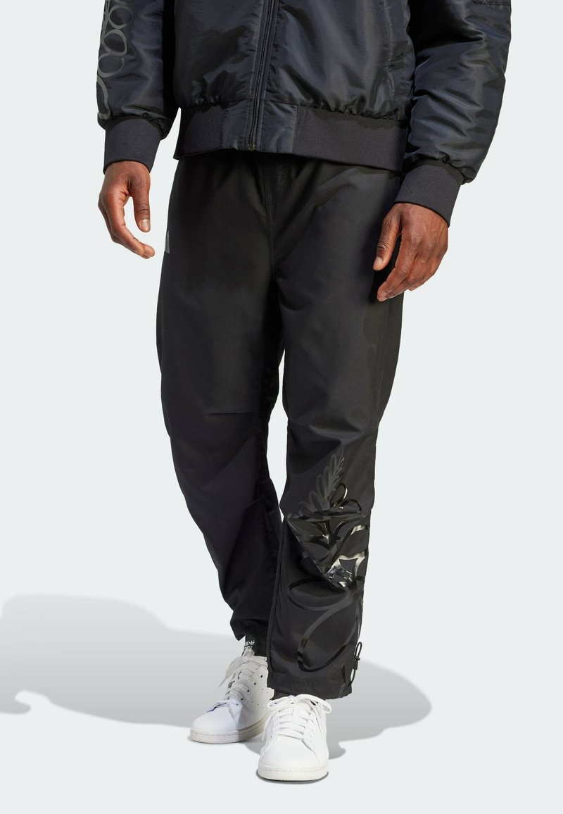 Personne portant un pantalon de jogging noir avec un motif noir brillant sur la jambe droite, une veste noire et des baskets blanches, debout sur un fond clair.