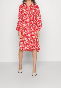 Robe florale rouge à manches longues, avec un boutonnière à l'avant et des poignets froncés ; tissu léger avec un fond crème ; portée avec des chaussures à talons carrés beiges.