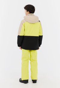 Kleurrijke kinderski-outfit met een hooded jack in zwart, beige en neon geel, gecombineerd met bijpassende neon gele broek en zwarte schoenen.
