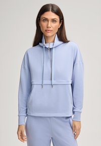 Donna con capelli castani lisci indossa una felpa con zip azzurra chiara e pantaloni abbinati, in piedi davanti a uno sfondo chiaro e uniforme.