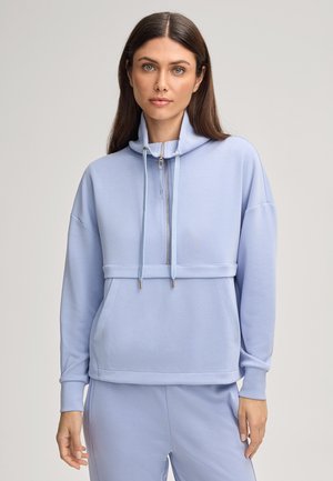 Frau mit glattem braunem Haar trägt einen hellblauen Reißverschluss-Hoodie und passende Hose, steht vor einem schlichten hellen Hintergrund.