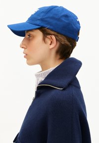 Junge Person im Profil, die eine blaue Baseballmütze, einen navyblauen Reißverschluss-Pullover und einen weißen Strick-Stehkragenpullover trägt, vor einem schlichten Hintergrund.