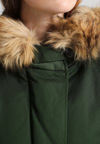Grøn vinterjakke med en brun faux fur krave. Stoffet har en glat tekstur med synlige søm og en knapdetalje.