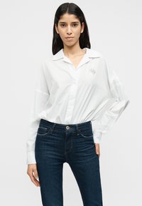 Λευκό πουκάμισο με κουμπιά, γιακά και μακριά μανίκια, με διακριτική κεντητή λεπτομέρεια. Συνδυασμένο με σκουρόχρωμο τζιν παντελόνι.