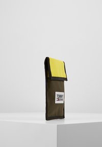 Portafoglio di tessuto verde con una pattina gialla e rifiniture nere. Presenta un'etichetta bianca con la scritta "TOMMY JEANS" sul davanti.