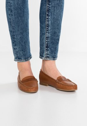 Person iført blå denimjeans og brune lædermoccasinloafers på et hvidt gulv med fødderne tæt sammen.