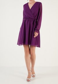 Žena v purpurových zavinovacích šatech s dlouhým rukávem a délkou po kolena, s vázáním v pase a světlými stříbrnými sandály na vysokém podpatku.