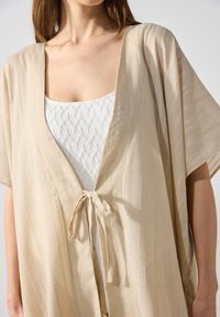 Beige kimono-tyylinen ylhäältä lyhyillä hihoilla, solmimisella kiinnityksellä, tekstuurikankaalla ja hienovaraisilla pystysuorilla raidoilla. Pukeutunut valkoisen istuvan yläosan päälle.