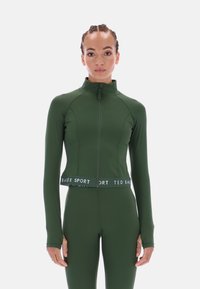 Zöld, rugalmas anyagból készült, cipzáras, rövidített dzseki, hosszú ujjakkal és márkajelzéses derékpánttal. Sima textúrájú és testhezálló szabású.