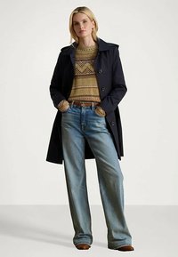 Mujer con un abrigo azul marino sobre un suéter beige con estampado, jeans de pierna ancha en un tono azul claro y zapatos marrones, de pie con las manos en los bolsillos.