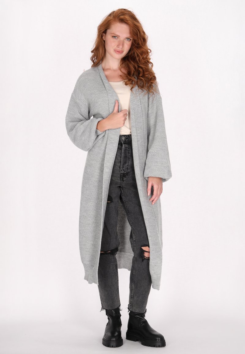 Lange grijze gebreide cardigan met een gestructureerd patroon, open voorkant en wijde mouwen, gecombineerd met zwarte gescheurde jeans en zwarte enkellaarsjes.