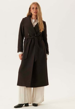 Vrouw in een lange zwarte jurk met riem over witte pantalon met wijde pijpen en zwarte hakken, staand tegen een effen lichte achtergrond.