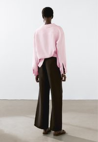 Světle růžová oversized košile s límečkem a bočními rozparky, spárovaná s černými širokými kalhotami a sandály, zobrazená zezadu.