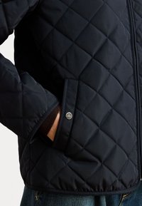 Veste matelassée bleu marine avec une texture lisse, dotée d'une poche latérale et d'un détail en bouton métallique sur le poignet.