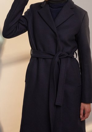 Personne portant un manteau bleu marine foncé ceinturé, avec des manches longues et un col, debout devant un fond neutre.