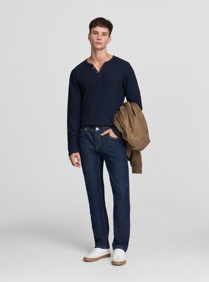 Giovane uomo con camicia a maniche lunghe blu scuro, jeans blu, sneakers bianche, che tiene una giacca marrone, in piedi davanti a uno sfondo chiaro e semplice.