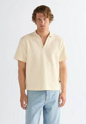 Polo color crema in tessuto strutturato, con scollo a V, maniche corte e colletto a contrasto. Abbinata a jeans azzurri.