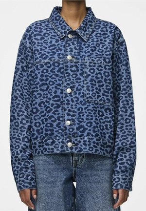 Personne portant une veste en jean imprimé léopard bleu avec des boutons argentés et un jean en denim bleu, debout devant un fond uni.