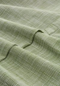 Tissu texturé en vert clair avec un motif à carreaux ; les plis créent des profondeurs et des ombres variées, mettant en valeur le détail tissé et un léger éclat.