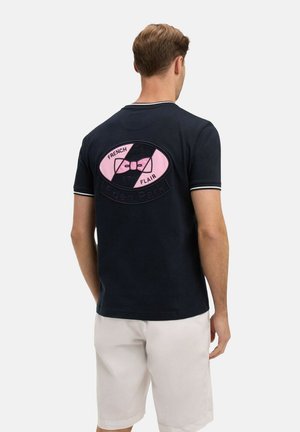 Homme portant un t-shirt bleu marine avec un logo nœud papillon rose et noir et le texte "French Flair" au dos, accompagné d'un short blanc.