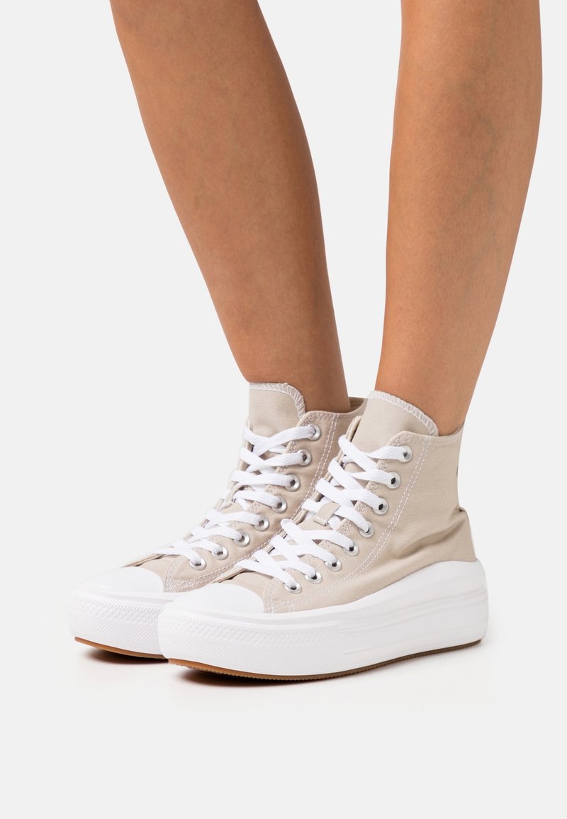 Jambes portant des baskets montantes beiges avec des semelles blanches épaisses et des lacets blancs sur un fond clair uni.