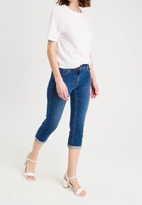 T-shirt blanc en coton associé à un jean capri bleu en denim, avec un ourlet roulé, complété par des sandales blanches à talons blocs.