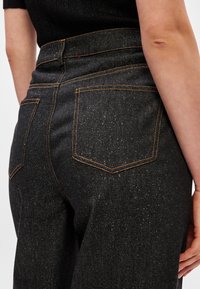 Calças de denim pretas com uma aparência texturizada subtil. Apresentam costuras laranjas e dois bolsos traseiros com um corte clássico.