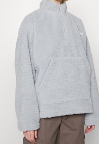 Hellgrauer Pullover-Jacke aus flauschigem Fleece. Verfügt über einen hohen Kragen, einen Viertel-Reißverschluss und eine große Fronttasche. Weite Ärmel.