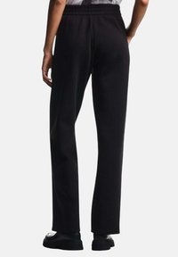Pantaloni sportivi neri con cintura elastica, gambe dritte e una texture liscia, abbinati a scarpe basse. La vista posteriore mostra le cuciture laterali.