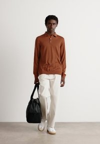 Mann in braunem langärmligen Polohemd, weißen Hosen, weißen Sneakers, der eine schwarze Reisetasche trägt, geht auf einem schlichten Boden vor einer weißen Wand.