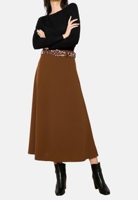 Bruine midilange rok van zachte stof, gecombineerd met een zwarte top met lange mouwen en een riem met luipaardpatroon. Zwarte enkellaarsjes met hak.