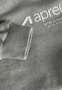 Harmaa collegepuseron resori kankaalla, jossa näkyy osa valkoista "aprel"-tekstiä ja maantieteellisiä koordinaatteja, jotka on painettu rintakehän alueelle.