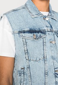Jasnoniebieska kamizelka dżinsowa z postrzępionymi otworami na ramiona, srebrnymi napami oraz dwiema kieszeniami na piersiach. Charakteryzuje się widocznym przeszyciem i klasycznym kołnierzykiem.