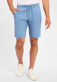 Helles blaues Shorts aus einem strukturierten Stoff, mit einem Kordelzugbund, Seitentaschen und umgeschlagenen Säumen. Getragen mit weißen Turnschuhen.